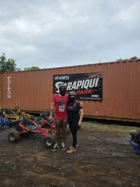 Dos visitantes con casco puesto junto a los go karts, frente al rótulo de entrada de Sarapiquí Race Park